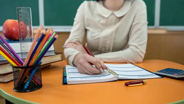 An educator sits at a desk with book, pencil, notepad and apple. Behind them is a classroom blackboard.