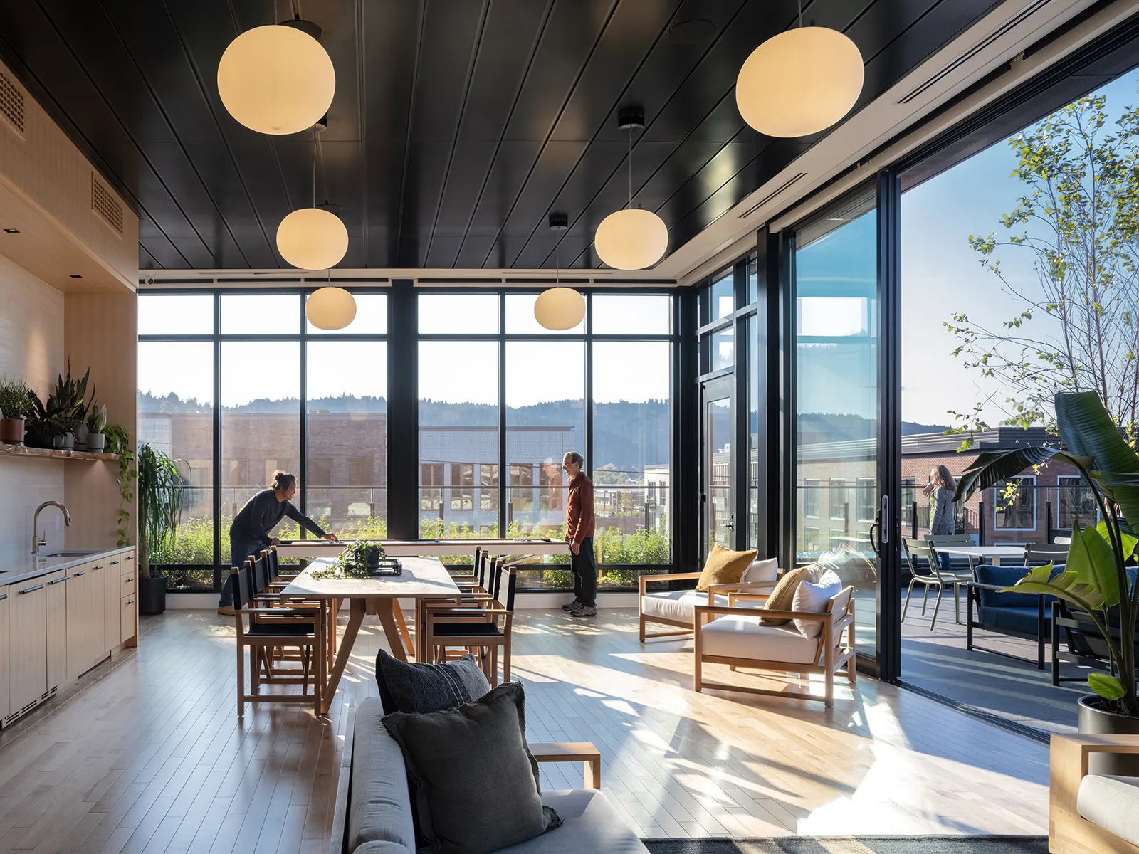 A room with many couches, tables, and chairs, along with glass walls looking out onto a rooftop.