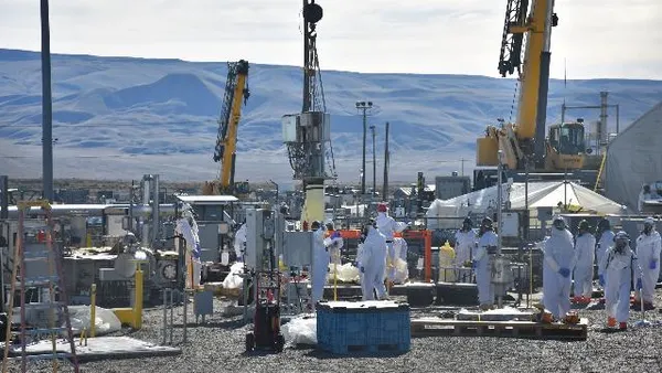 A group of people in white hazmat suits move about on an industrial site.
