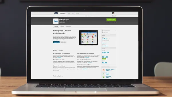 A laptop sits on a wooden desk displaying a webpage for “Box OneCloud – Enterprise Content Collaboration.” The screen shows product information, pricing, and a tablet image with app icons. The webpage includes blue buttons labeled “Take the Tour” and “Watch Demo,” along with a green “Try the Free Edition” button at the top right. The background is softly blurred with a dark wall and a framed pictu
