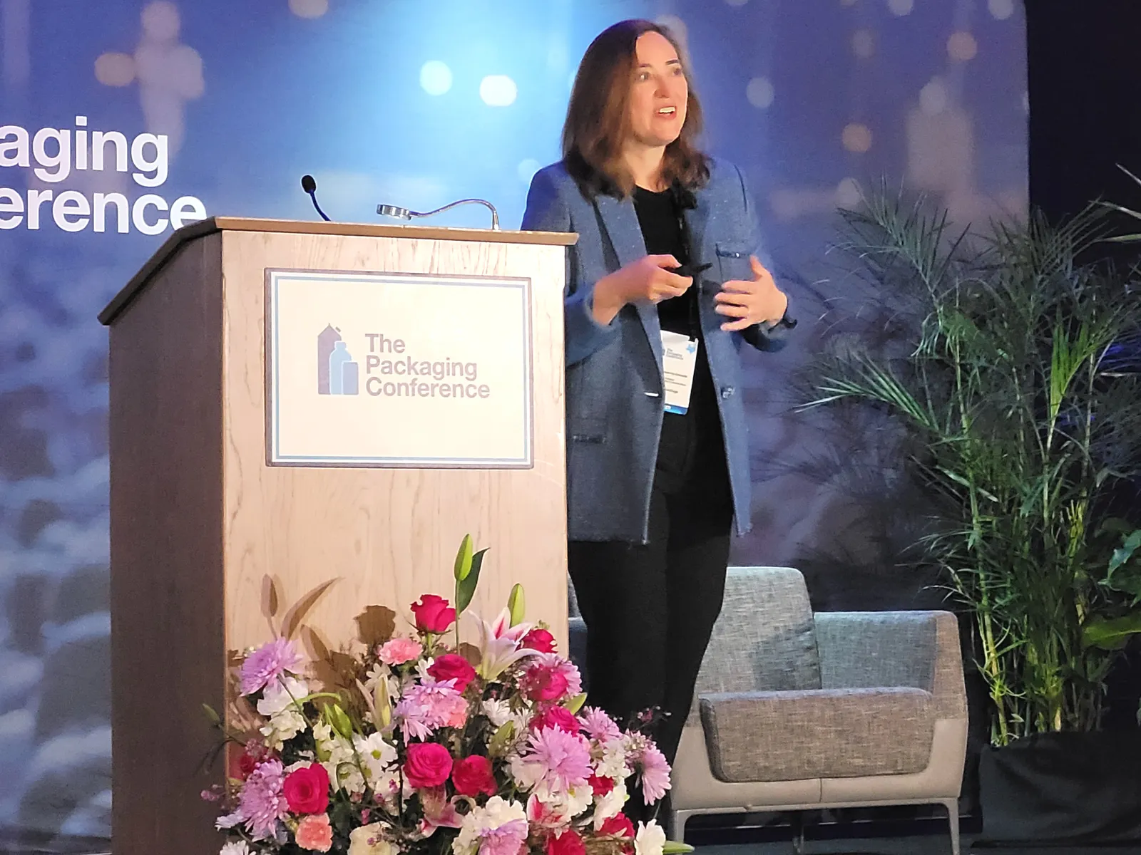 A person stands on stage next to a podium with branding for The Packaging Conference.