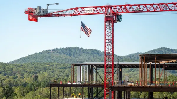A crane looms above a steel structure of a building. In the background are large, verdant trees.