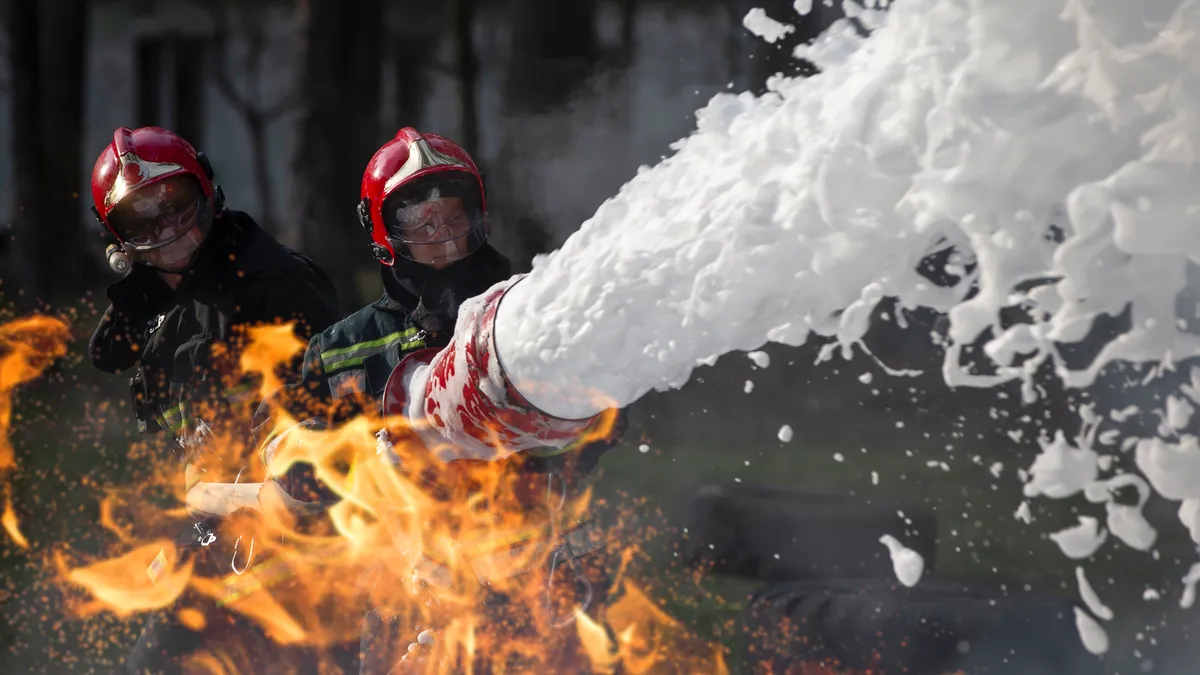 Firefighters in red, black and yellow protective gear spraying white aqueous film-forming foam to extinguish a fire.