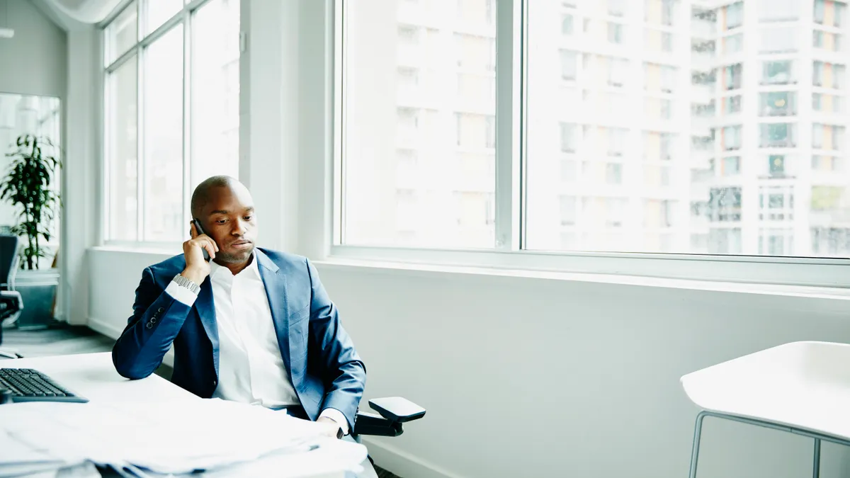 Frustrated businessman sitting at office workstation waiting while on smartphone