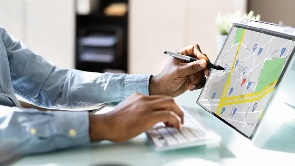 A person's uses a computer keyboard with one hand and points to a map on a screen with the other.
