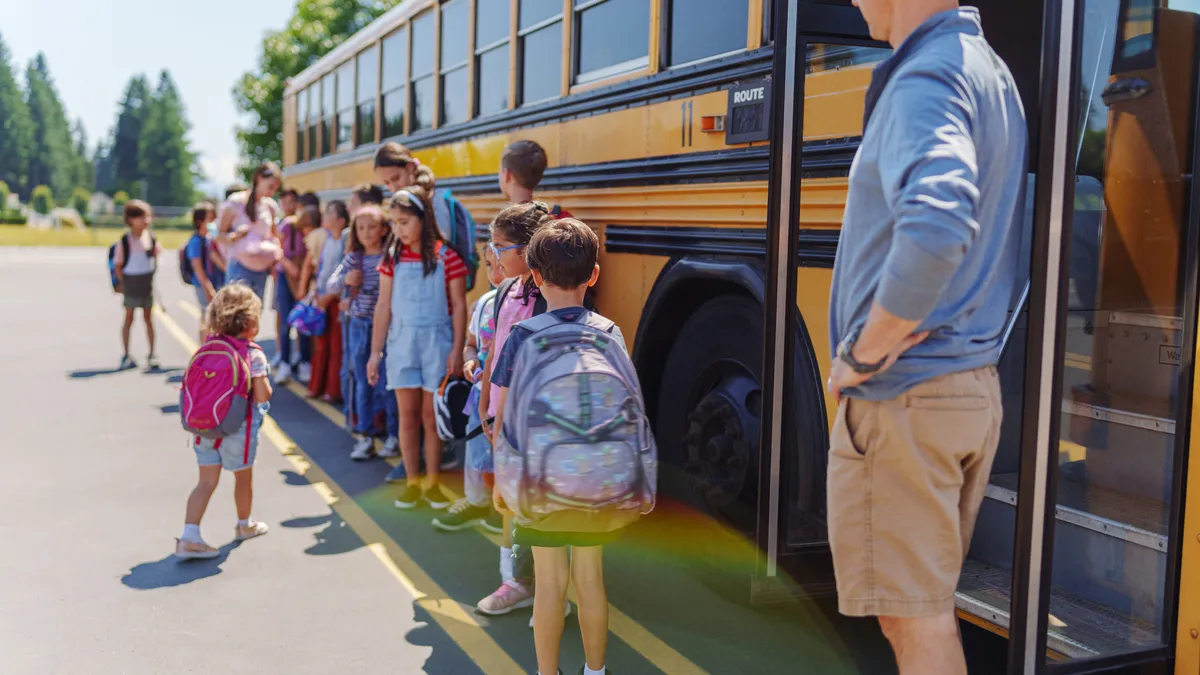 A group of young elementary-age children line up outside a yellow school bus with backpacks on as an adult man looks over them