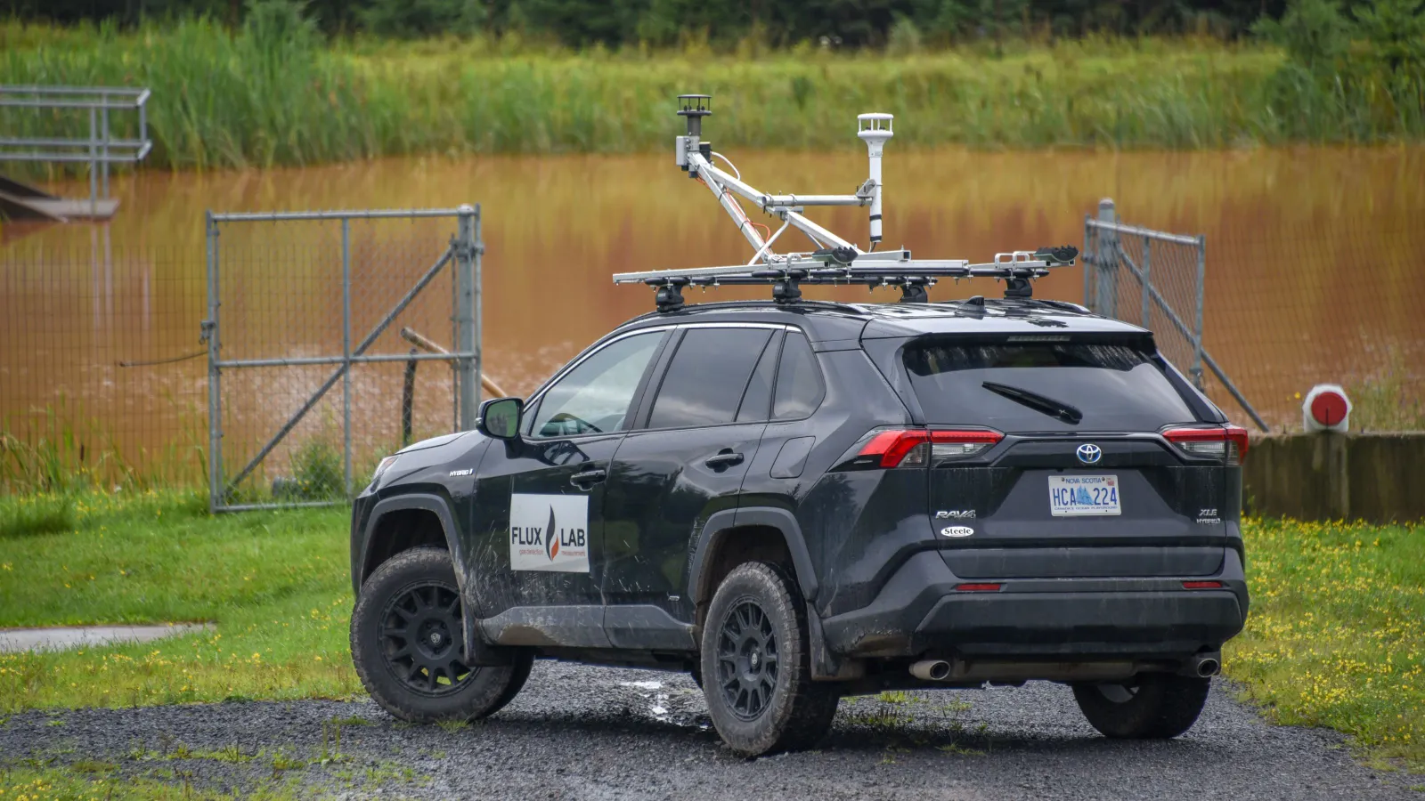 An SUV with the FluxLab logo on its side and scientific instruments mounted to its roof rack with brackets is parked on a gravel road. Behind it is a fence, creek and wooded area.