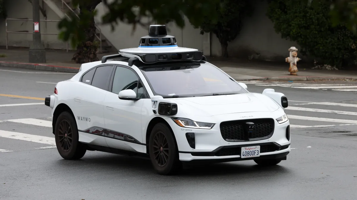 A white sedan with large sensors mounted on the roof and front hood drives along a street in San Francisco.