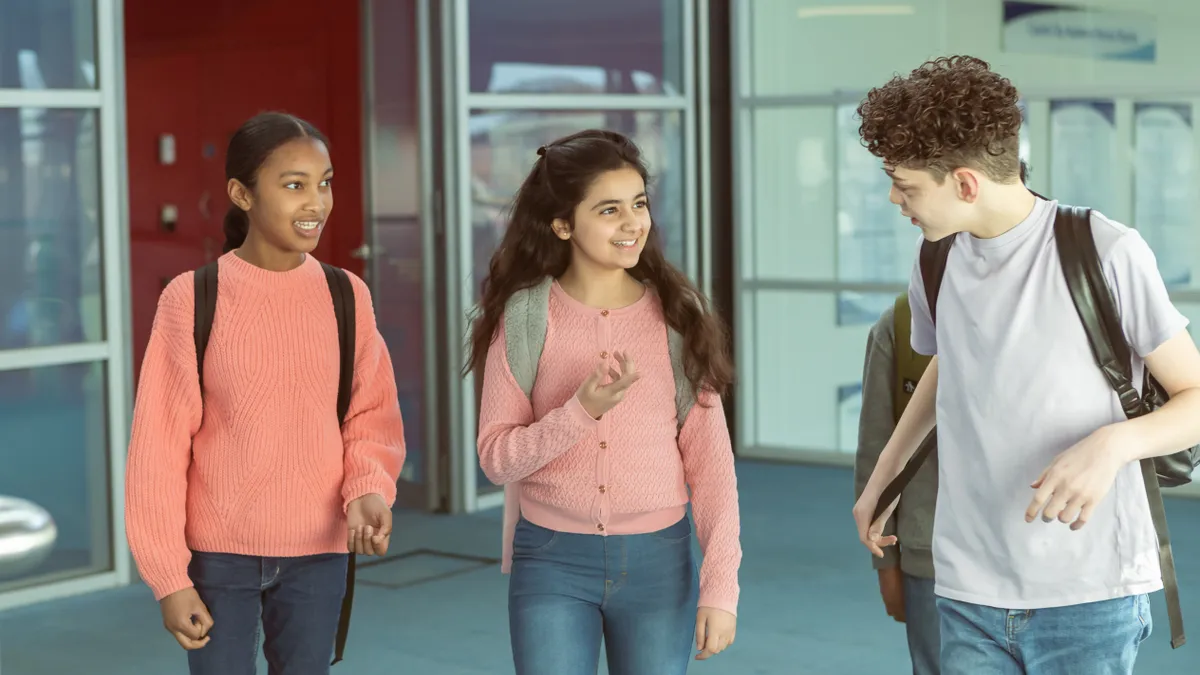 Three middle school students walk in a hallway of a school building. They are looking at each other and carrying backpacks.