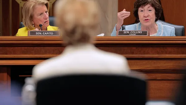 Two people are seated at a dias in a hearing room. They are facing the camera and looking at one person seated at a desk whose back is to the camera.
