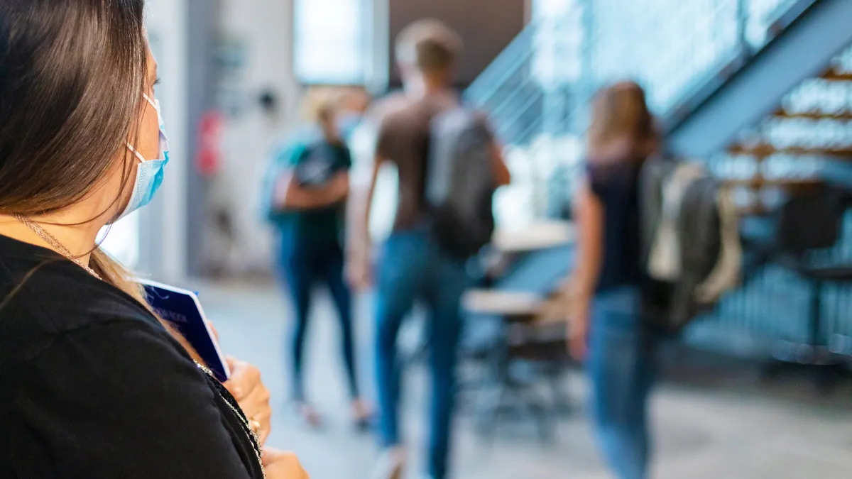 A teacher wearing a face mask looks out into a hallway of high school students walking away while wearing their backpacks.