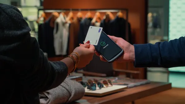 A customer taps a Visa card on a smartphone payment terminal inside a clothing store, with the phone screen showing an ‘Approved’ message.