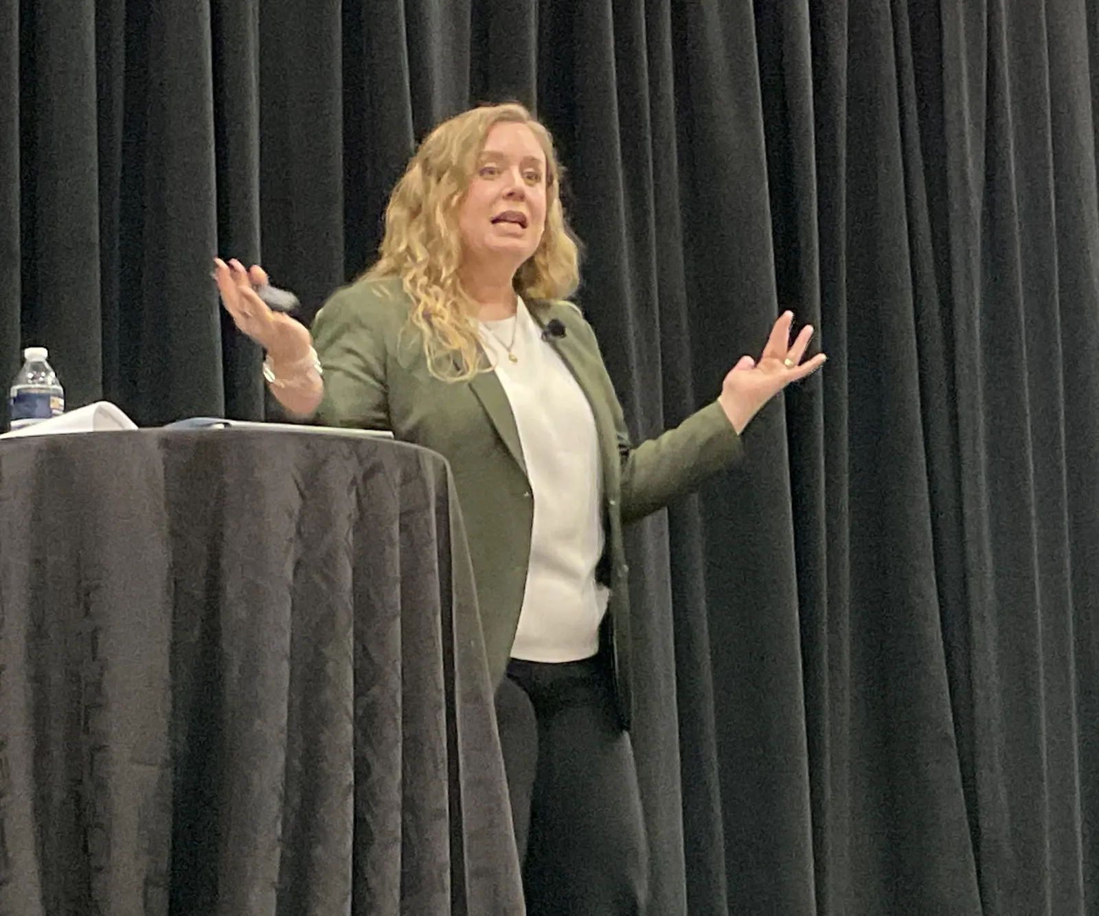 A woman speaks while standing next to a table in front of a black drape