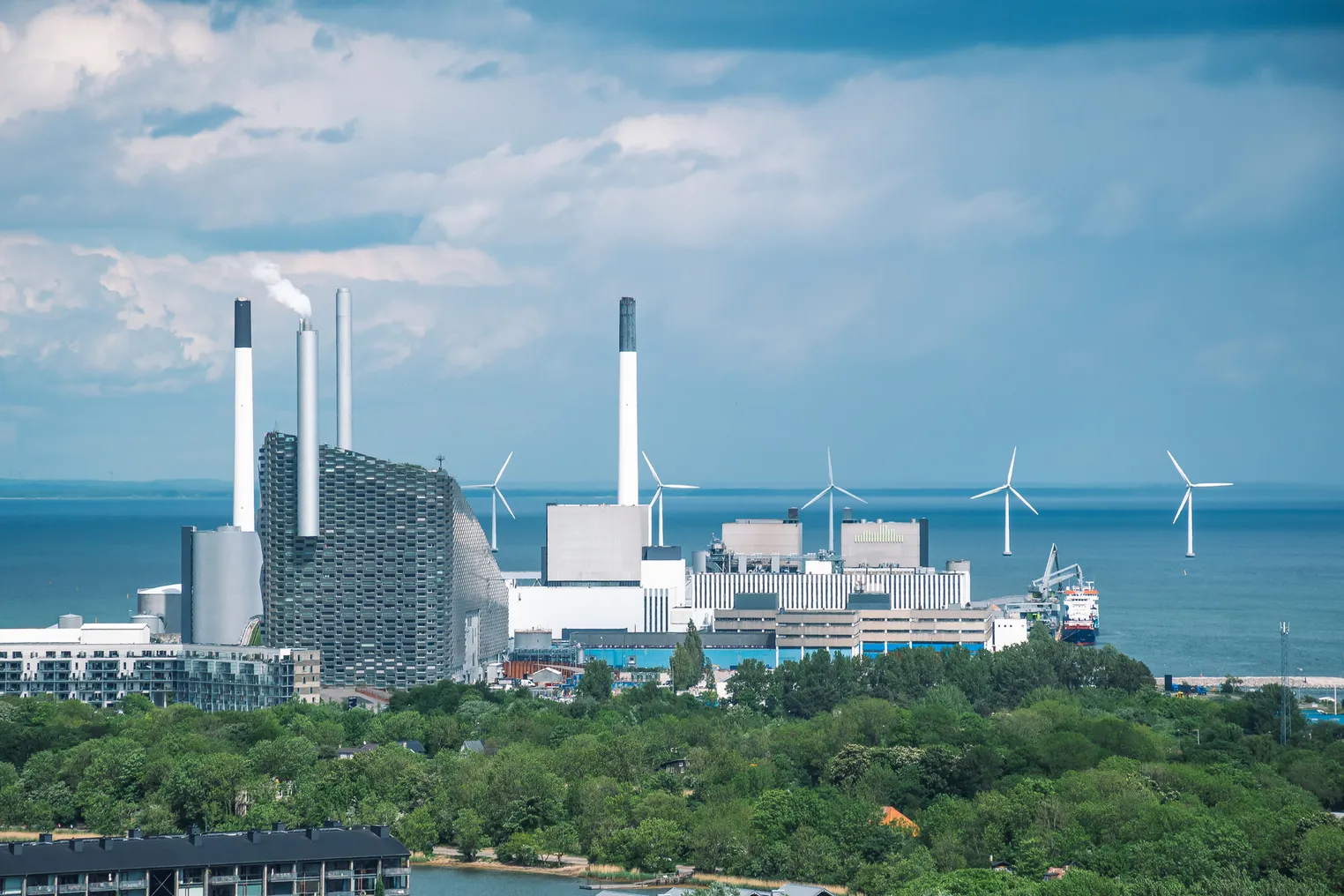 Industrial waste plant with smokestacks in Copenhagen, alongside waterfront with wind turbines in the background