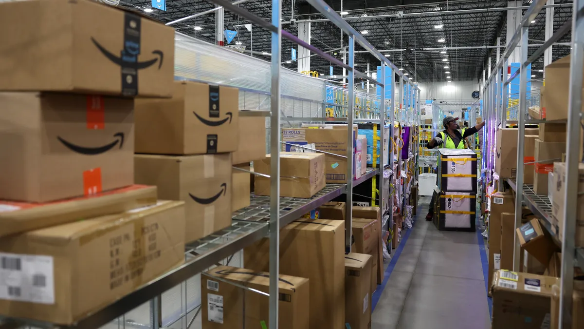 An Amazon worker with a cart sorts packages in a warehouse aisle packed with Amazon packages.