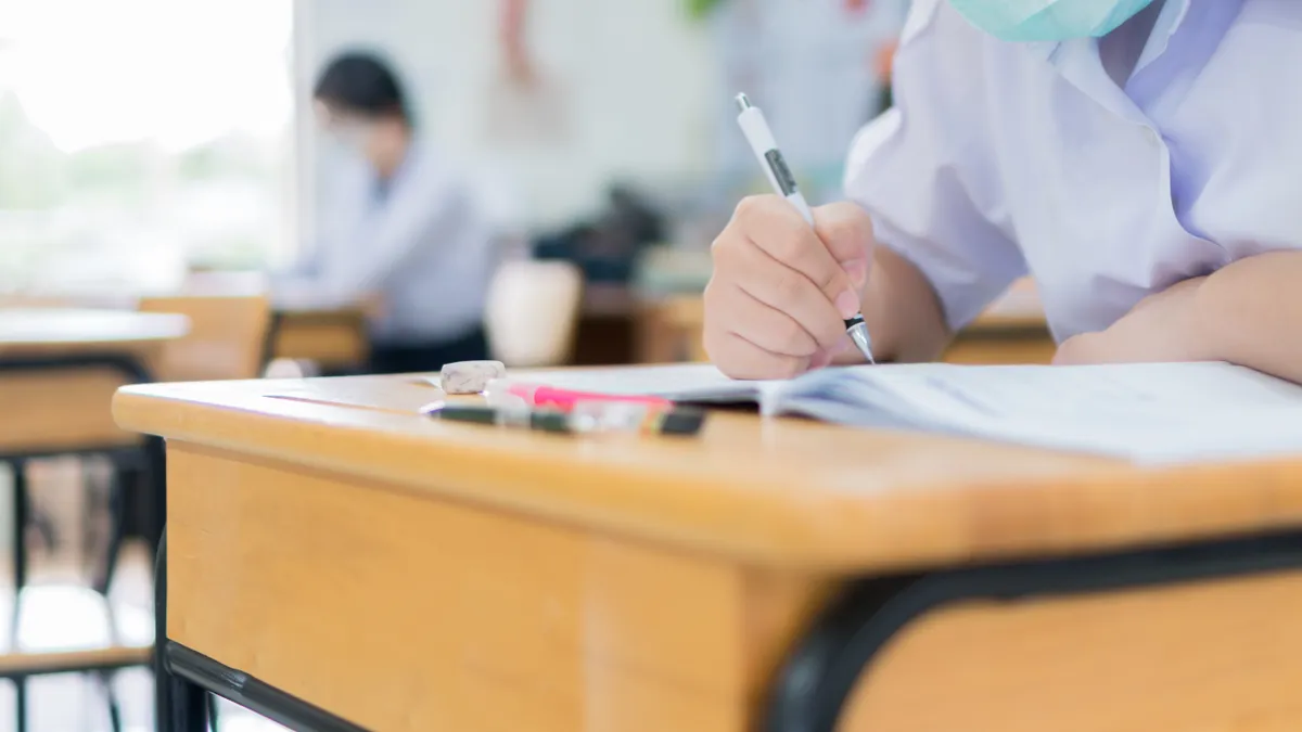 A young student wearing a face mask writes with a pen while working in a classroom.