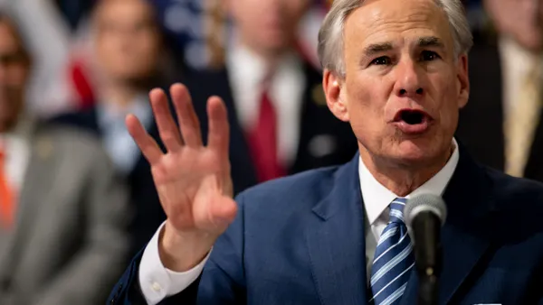 A man gestures before an out-of-focus background of people gathered.