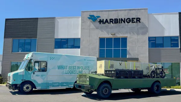 A white and light turquoise delivery truck with the Harbinger sign imprinted on it, parked next to a green military truck.