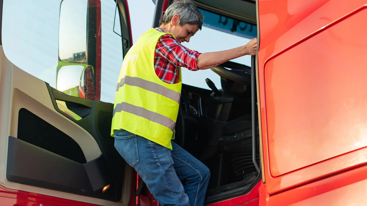 A woman truck driver hangs to the side of a cab by the driver's seat as she enters the cab.