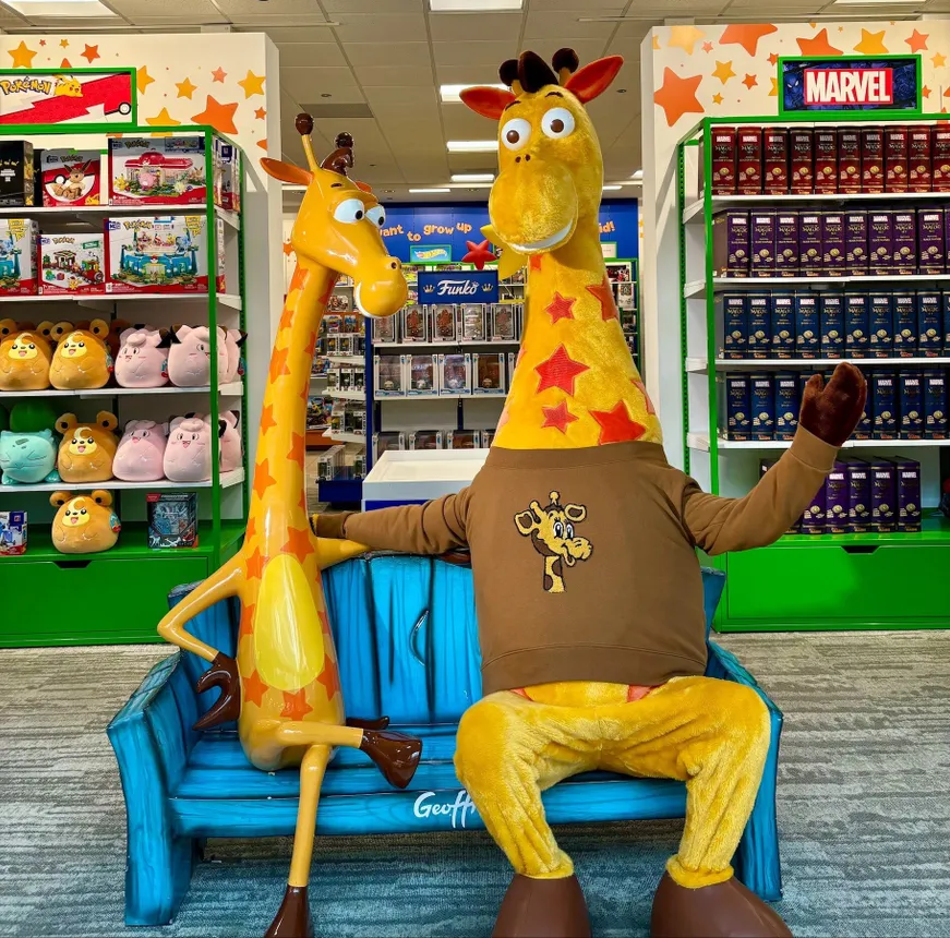 A Geoffrey the Giraffe mascot wearing a sweater form Bonobos&#x27; collaboration with Toys R Us, sitting next to a Geoffrey the Giraffe statue.