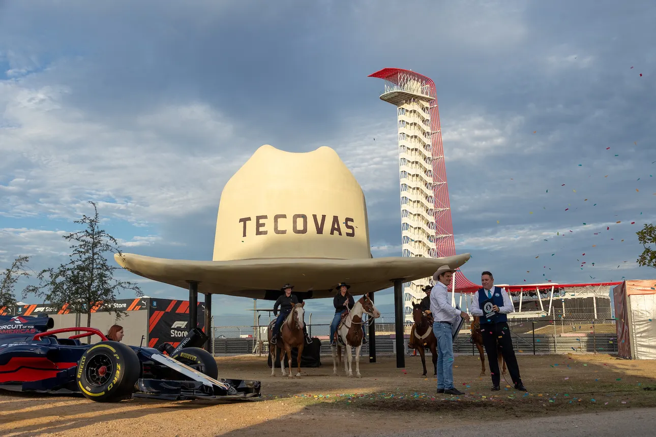 Two men riding horses under a giant cowboy hat, a race car in the foreground