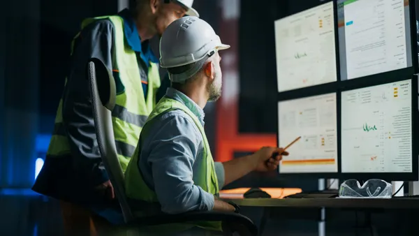 Two construction workers look at a series of computer monitors.