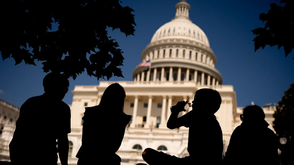 Trees and sitting people are silhouetted against a white government building and blue sky.