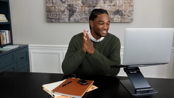 A person sits at a desk, smiling, with a square device on top of a stack of notebooks. They are looking at a laptop.