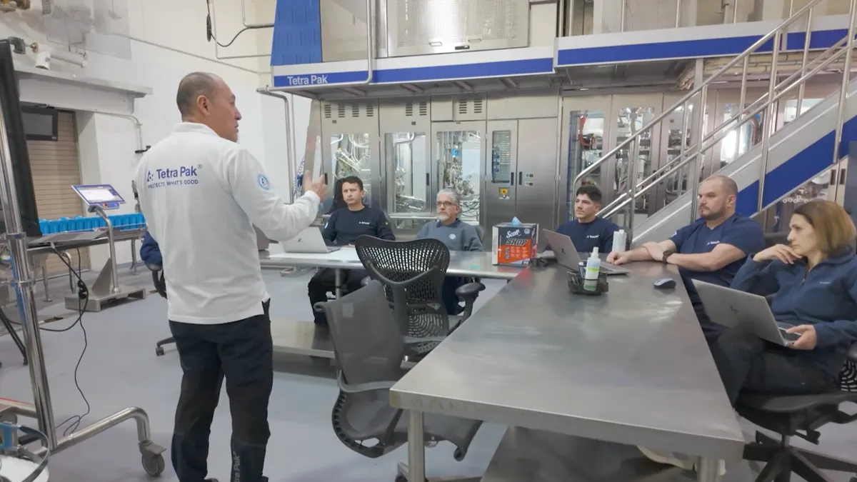 One person stands and speaks to five people seated at a metal table in an industrial setting with a Tetra Pak logo on the wall.