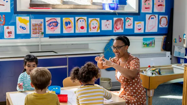An adult is sitting at a small table with young students in a classroom. The adult is making a motion with their hands and the students are watching. Paintings of faces are on the back wall.
