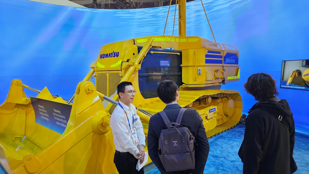 People stand in front of a large, yellow piece of heavy machinery in front of a light blue, underwater-like background.