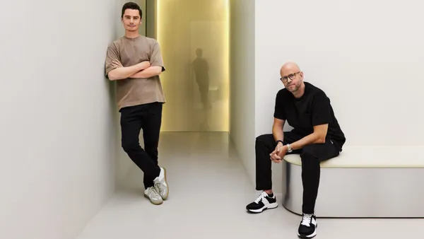 A picture of two people staring at the camera, one is leaning against a wall and one is sitting on a bench, in a white room.