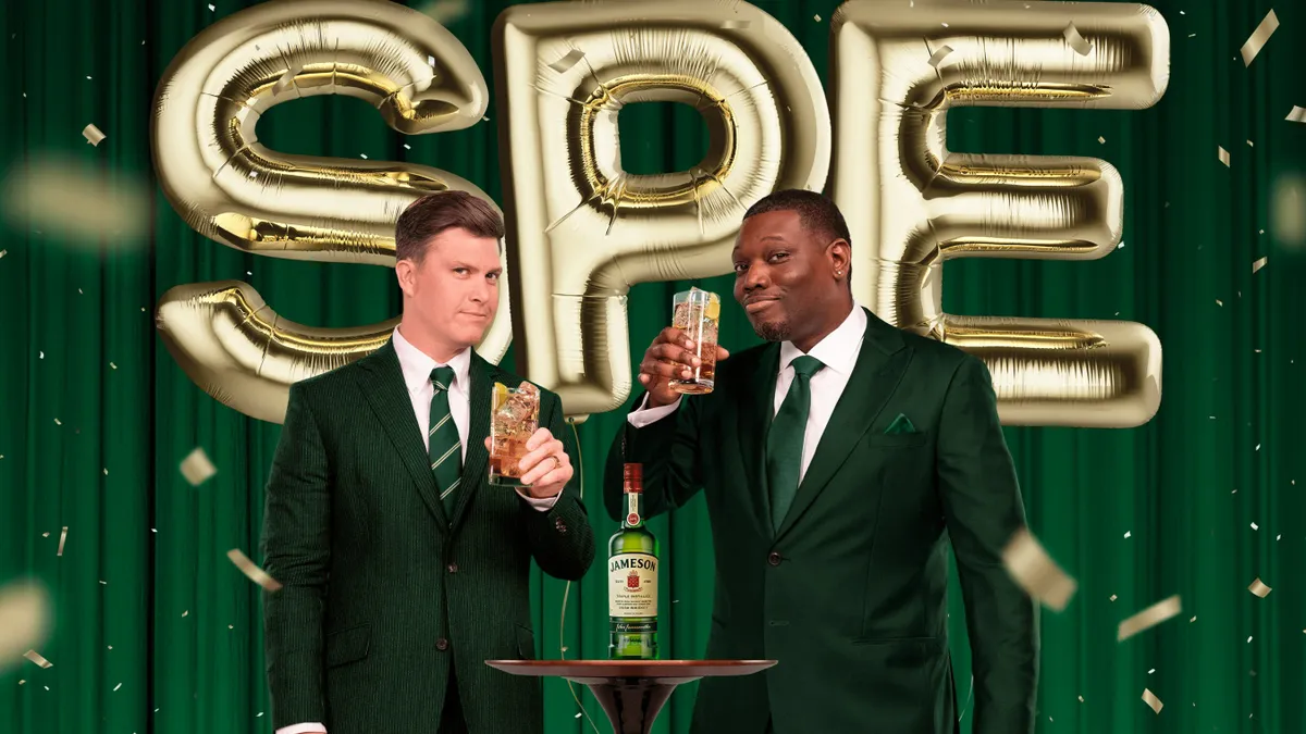 Colin Jost and Micheal Che wear green suits in front of a green background with gold balloons which spells out SPE. Both comedians hold up a cocktail with a bottle of Jameson between them.