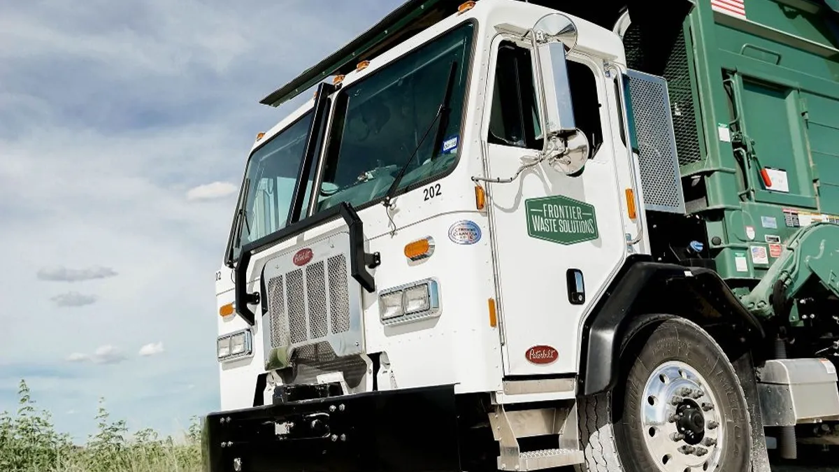 The cabin of a waste truck is featured prominently. It sits on a white gravel road with brush behind it.