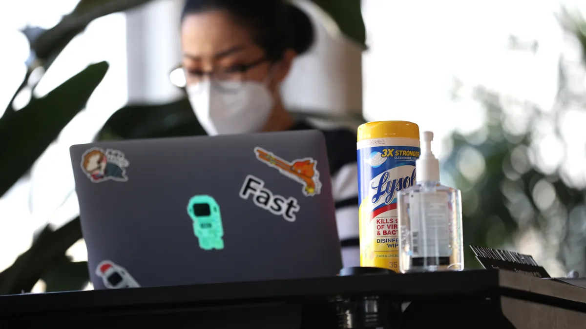 A worker wears a mask and sits at their desk, which contains disinfectant spray and wipes.