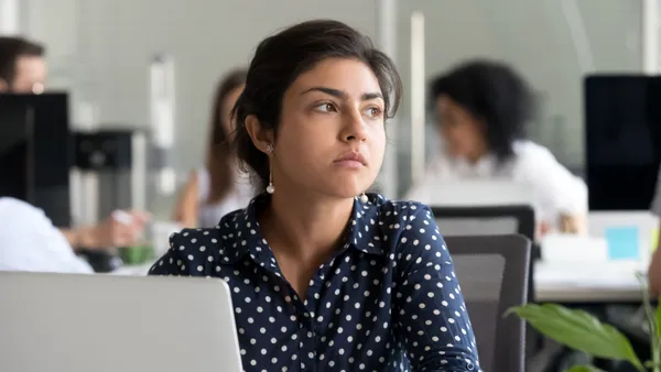 An office worker in a polka-dotted shirt sits at her computer and looks thoughtfully in the distance.