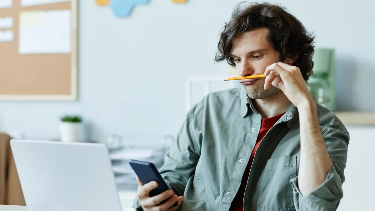 Portrait of Caucasian young man playing with pencil and using phone at workplace suffering from boredom and procrastination