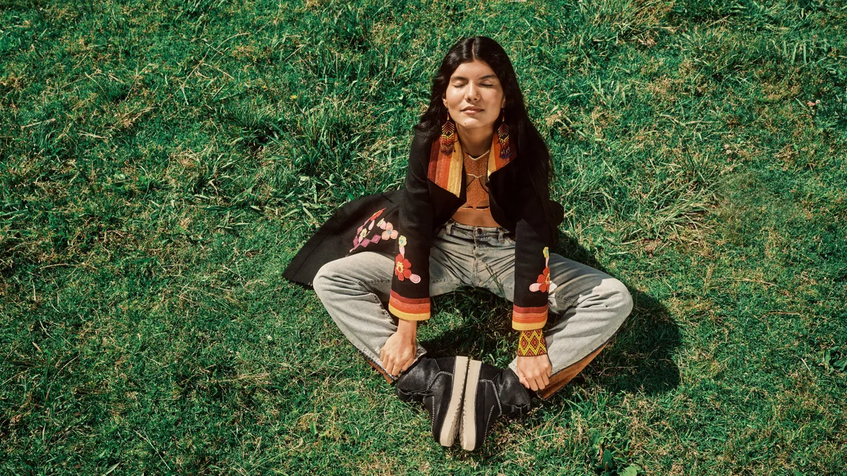 Zaya Guarani sits on a grassy field wearing a graphic black jacket, beige pants, and modeling a black pair of Campfire Crafted Regenerate Boots by Ugg.