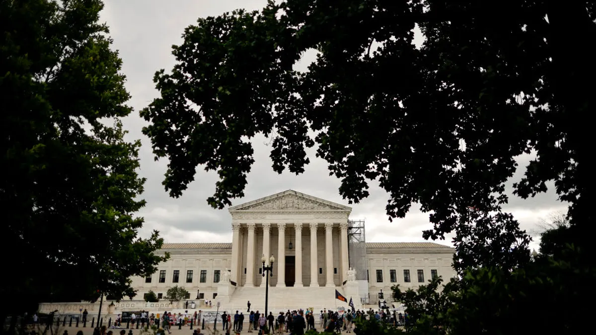 The facade of a large white building framed by trees is seen. People are gathered in front of the building.