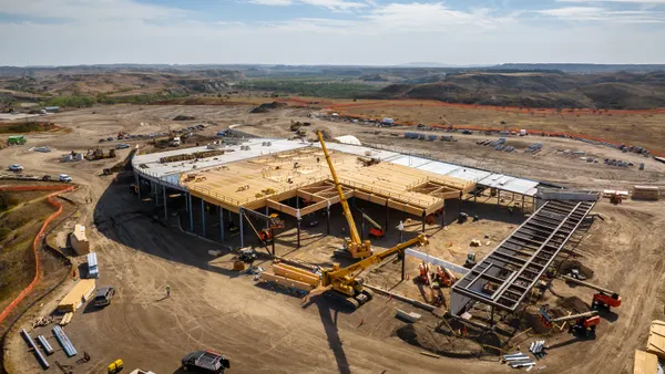 An overhead shot of a massive wooden building that stands on dirt in the middle of a bright day with clear skies.