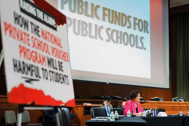 A person is seen sitting at a dias in the background in front of a large screen that says "Public Funds for Public Schools" Another sign is in the foreground