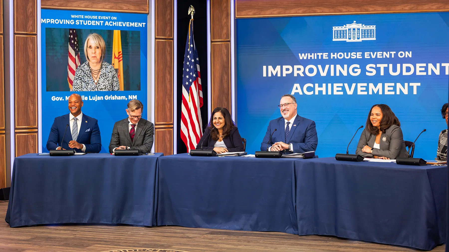 Six adults sit behind a long table. Behind them is a screen reading "White House Event on Improving Student Achievement." Another person is on a screen in the background