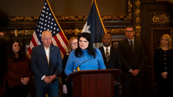 A person in a blue shirt speaks at a podium with people in suits and ties lined up behind her