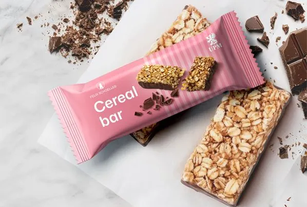 A snack bar with a pink wrapper lies on top of an oat bar next to pieces of chocolate.