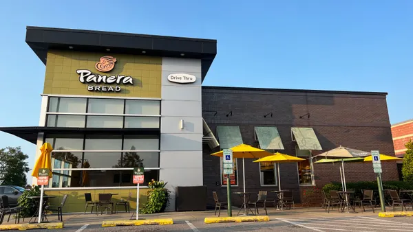 A photograph of a building that with white signage that says Panera Bread. There is an empty parking lot out front and an outdoor patio on the right.