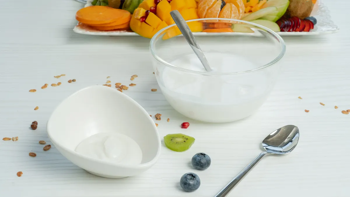 A small white dish of plain yogurt made from cell-cultured dairy sits in front of a larger glass bowl of the yogurt. A spoon lies in front and a cutting board of fruit is in the background.