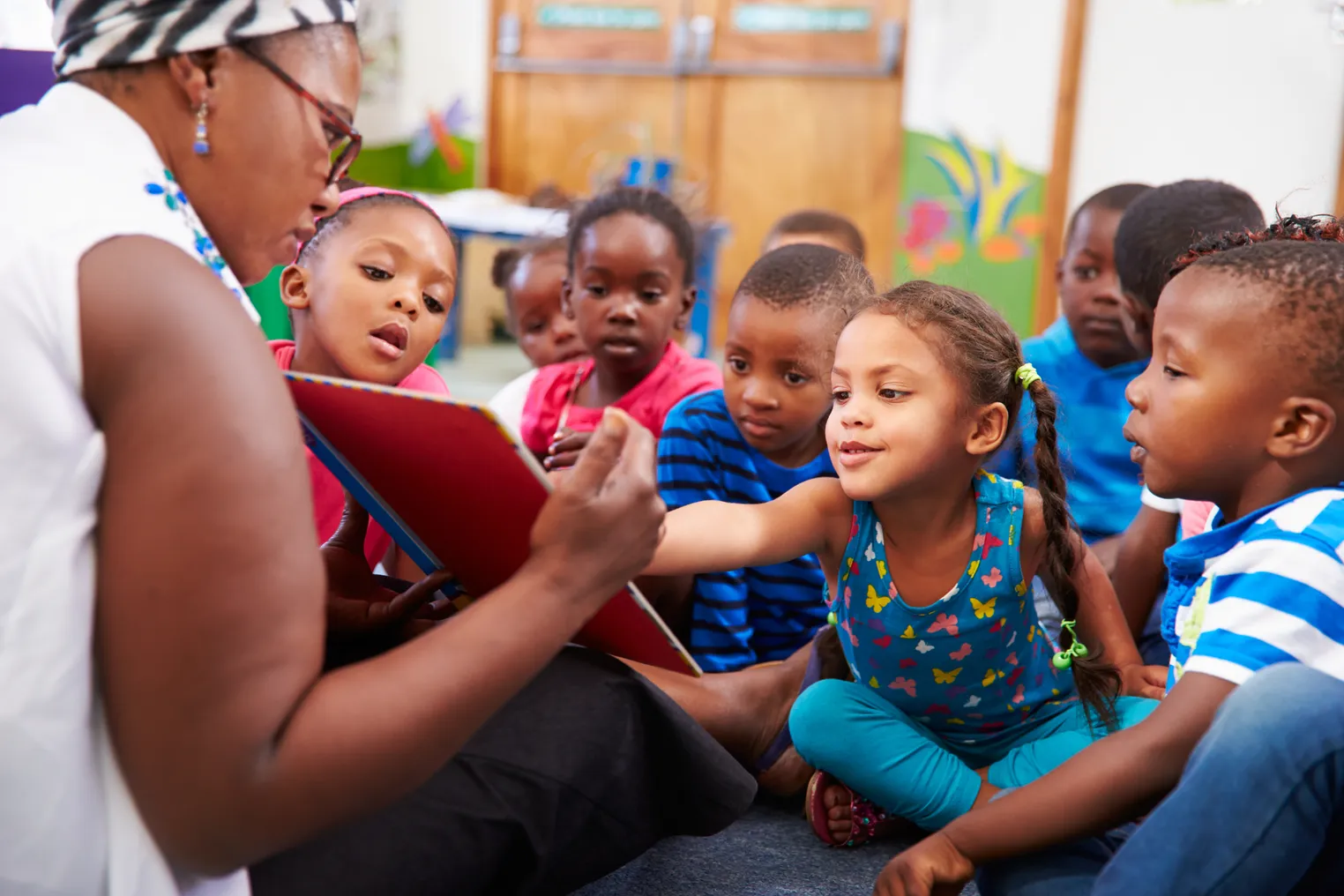 An adult sits on the floor holding a book with back toward camera. A group of young students sit facing the adult and look at the book.