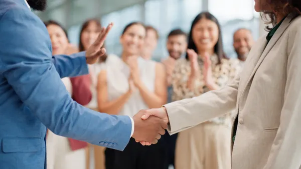 Two people in business attire shake hands, while others clap in the background.