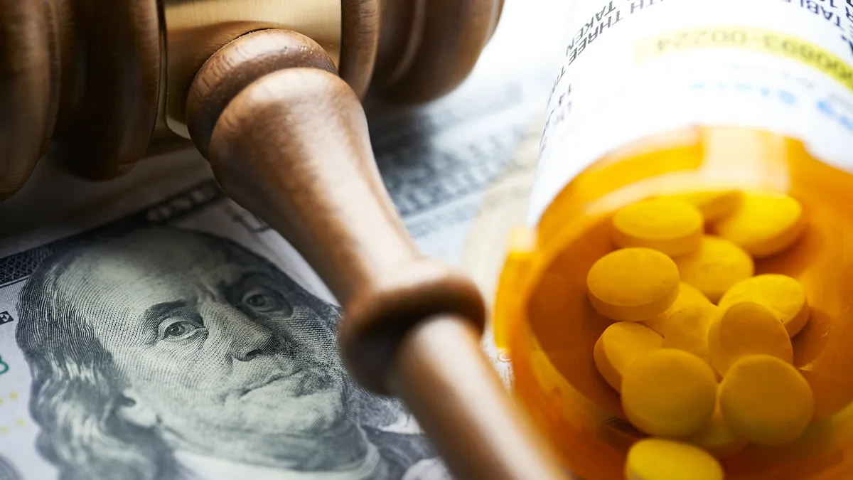 A prescription bottle with white pills and a wooden gavel sit atop a $100 bill showing Benjamin Franklin's face.