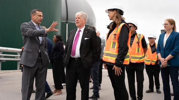 Two people in suits stand in front of large rounded buildings, while others wearing construction vests and hard hats accompany them.